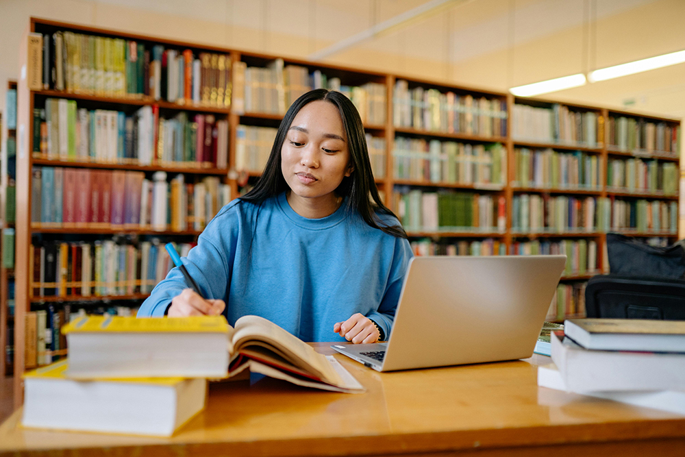 a person writes in a library in front of a laptop