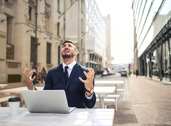 A frustrated man sitting outdoors in front of a laptop looks up at the sky in despair