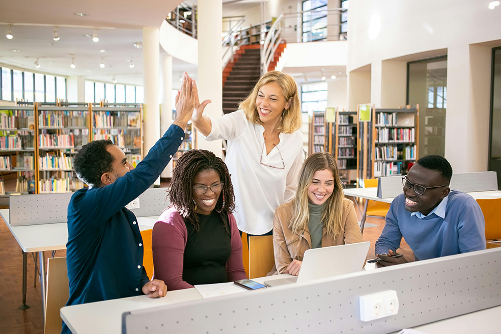 Five people sit and stand around a laptop in a library; two of the people high-five